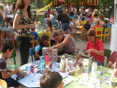 Copii și adulți la mese de lucru în aer liber, cu materiale de pictură și cutii reciclate