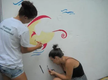 Două voluntare pictează un soare pe peretele unei grădinițe